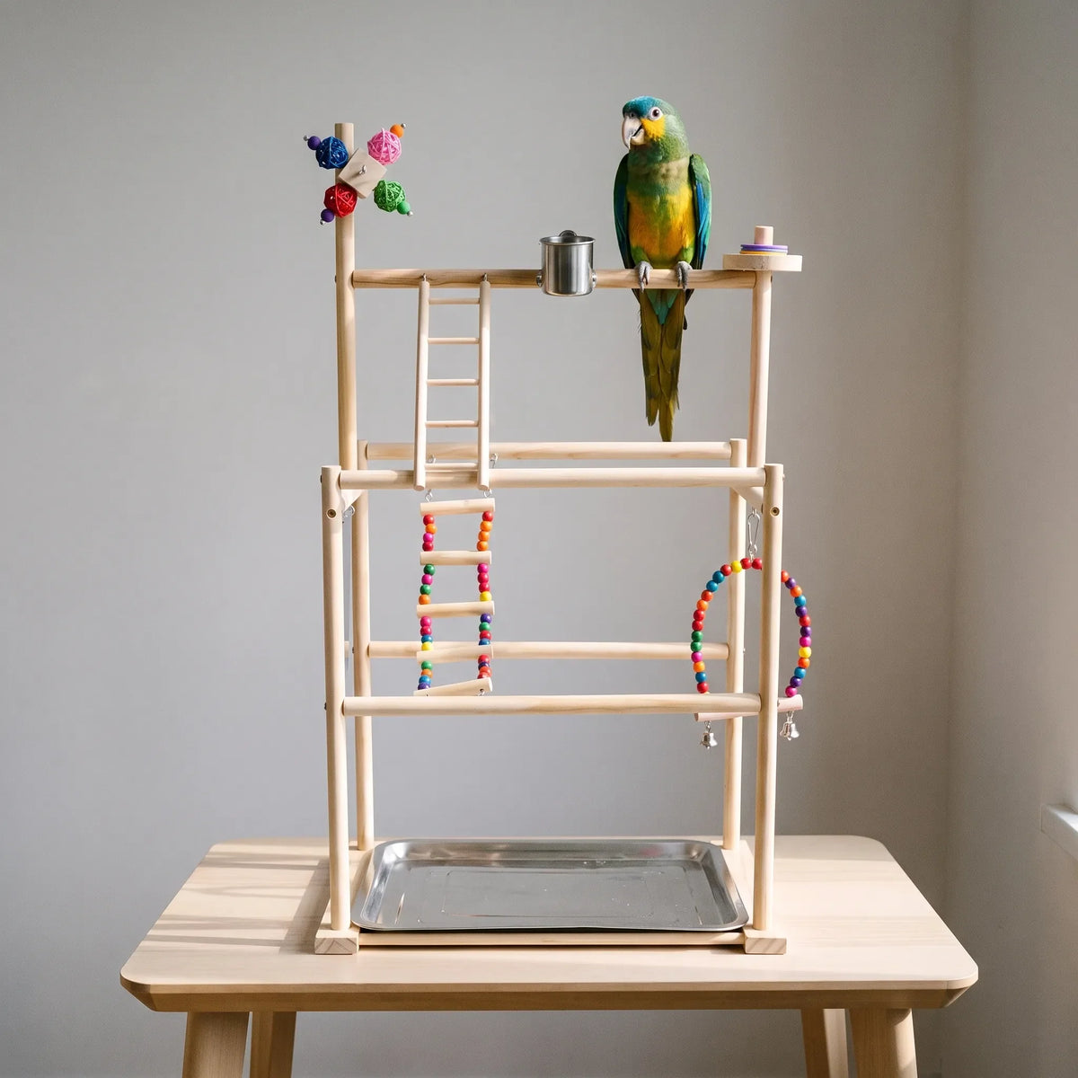 Wooden playstand on a pale table with a green and yellow parrot perched on the top bar beside the steel cup. 2