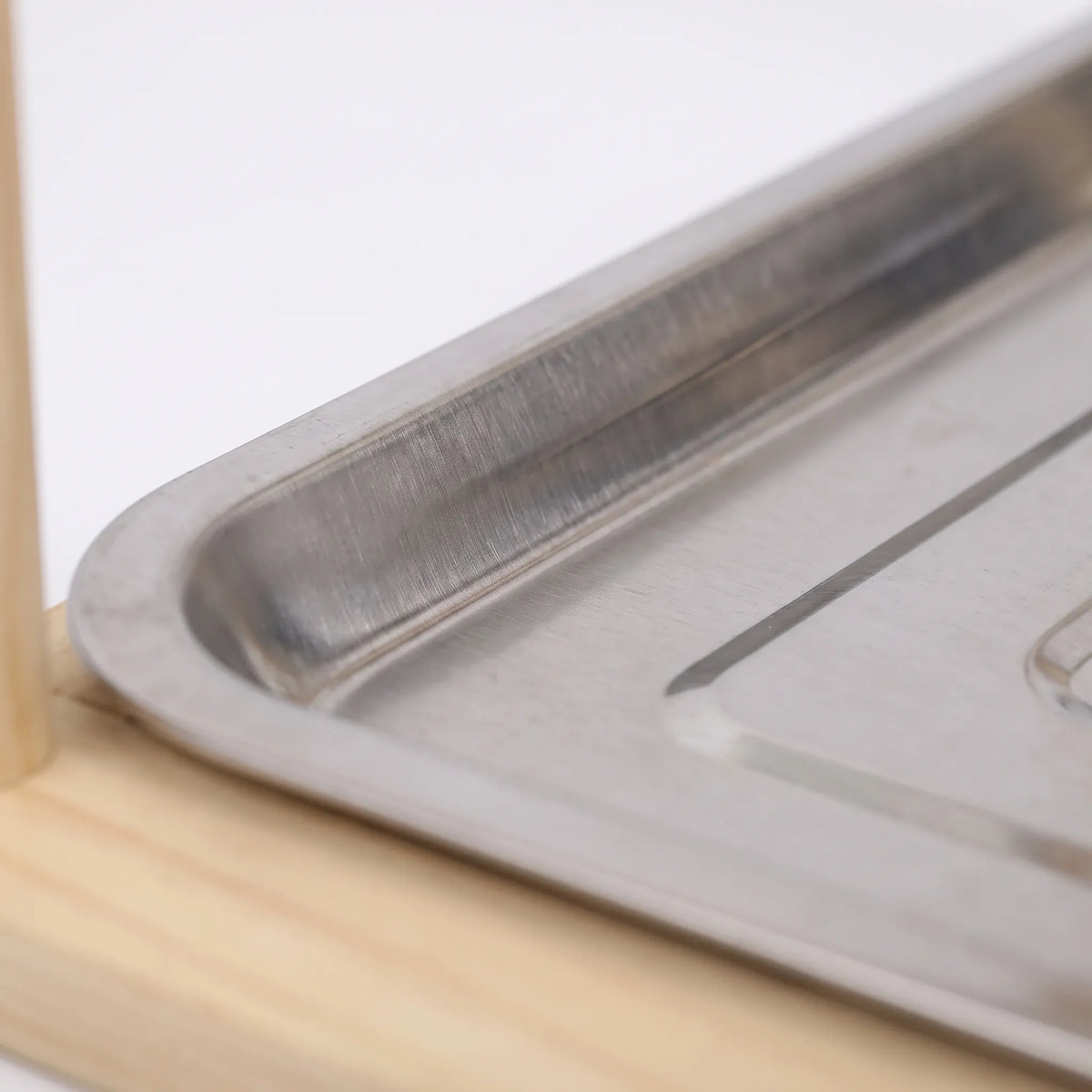 Close-up of the stainless steel droppings tray corner resting on the pale wooden base, with a post at left. 3