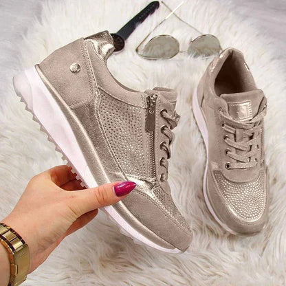 Hand holding one taupe trainer to show its side zip and white sole, with the other shoe, sunglasses and watch on a fluffy rug. 7