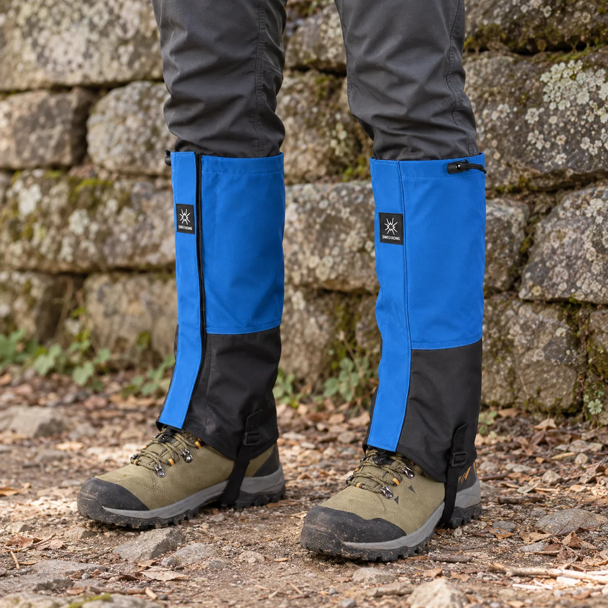 Blue and black gaiters with side zips worn over green hiking boots on a stony woodland path beside a lichen-covered wall. 1