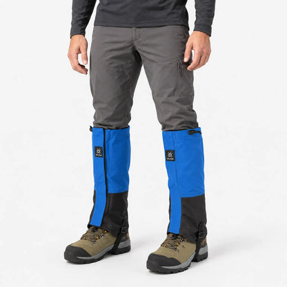 Man standing in a studio wearing blue and black gaiters over grey trousers and olive hiking boots, seen from the front. 2