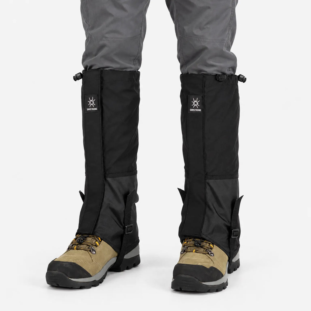 All-black gaiters with a two-tone panel and underfoot straps, worn over grey trousers and tan hiking boots on a white background. 8