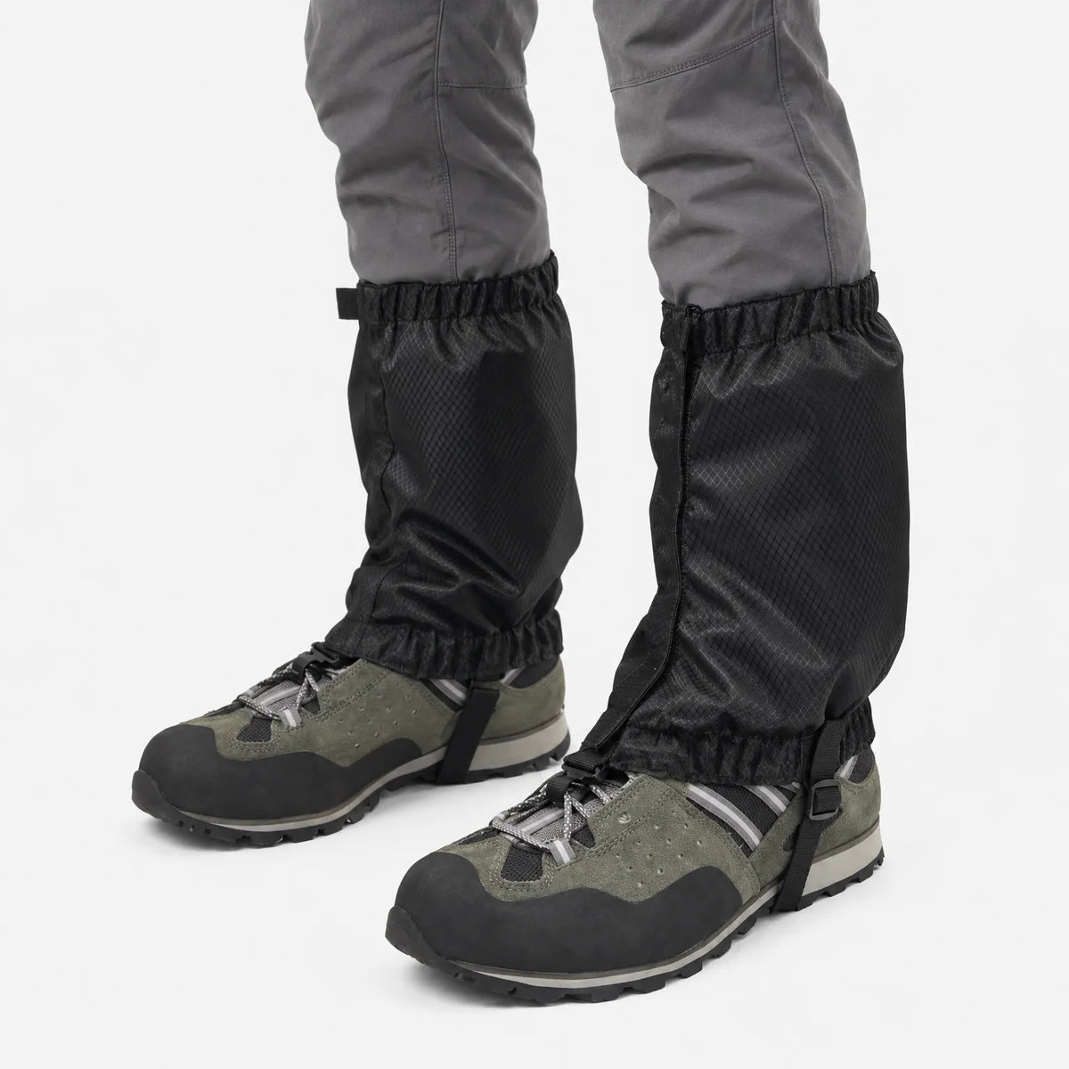 Black gaiters on grey trousers and olive hiking boots, angled view showing the side closure and underfoot strap, white background. 5