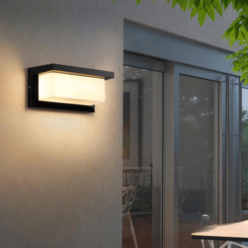 Black rectangular wall light glowing warmly on a rendered exterior wall beside glazed doors at dusk 1