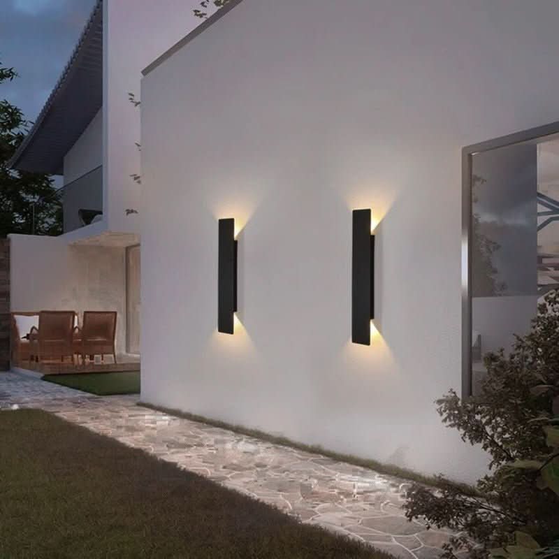 Pair of black wall lights on a white garden wall at dusk, throwing warm light up and down over a stone path. 3