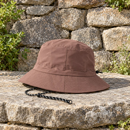 Brown bucket hat resting on a sunlit stone ledge, drawcord with toggle trailing beside it, dry-stone wall behind. 3