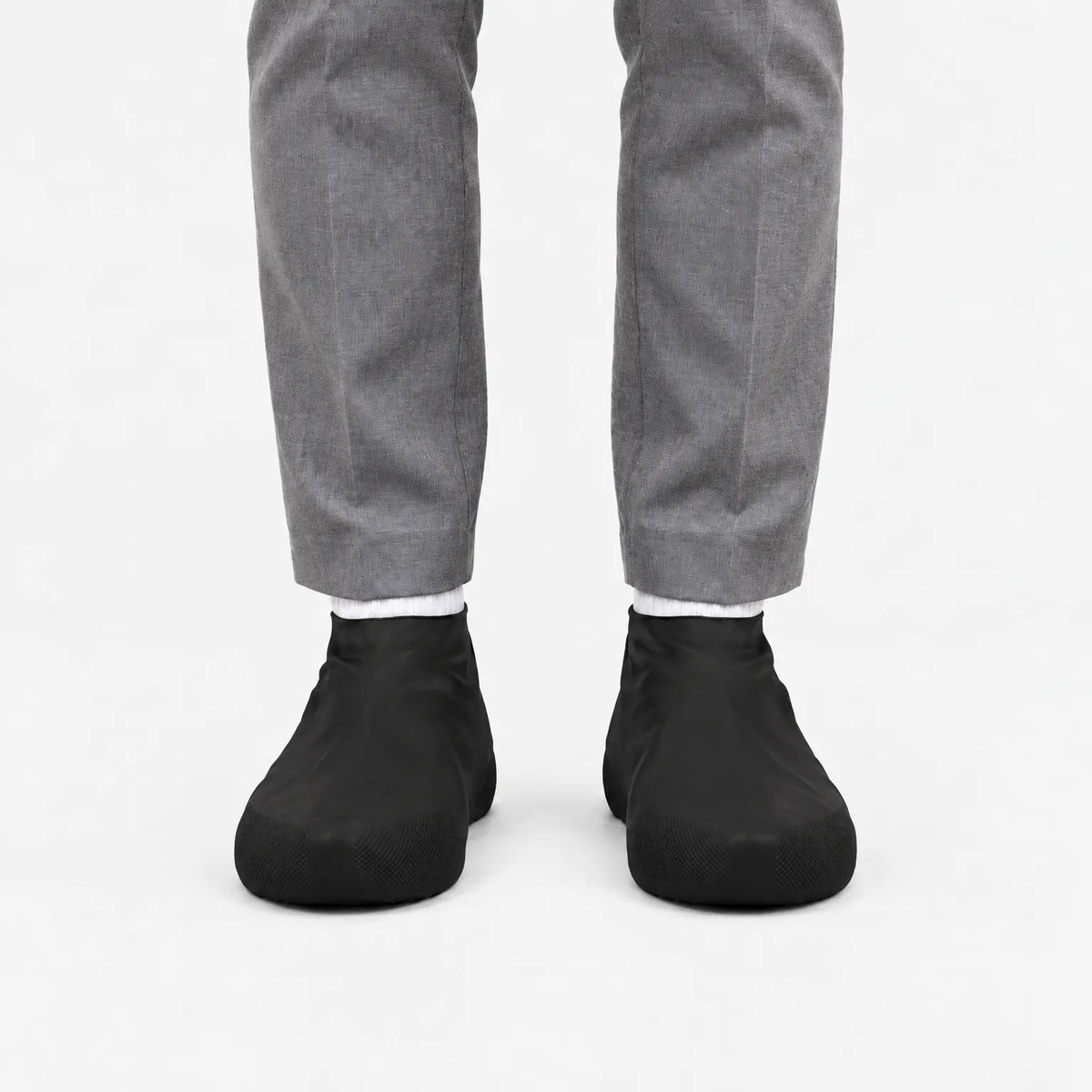 Front view of black shoe covers worn with white socks and grey trousers, with textured grip panels at the toes. 9
