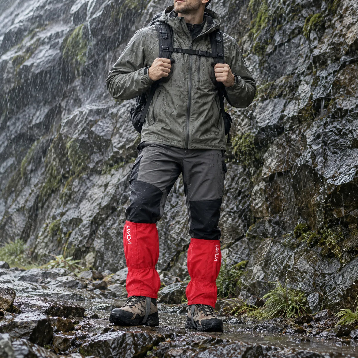 Hiker in red gaiters over walking boots stands beside a wet, mossy rock face in heavy rain, wearing a waterproof jacket and backpack. 1