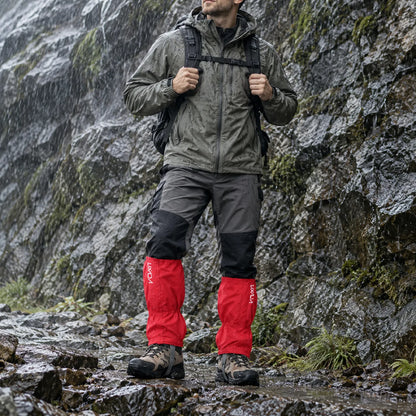 Hiker in red gaiters over walking boots stands beside a wet, mossy rock face in heavy rain, wearing a waterproof jacket and backpack. 1