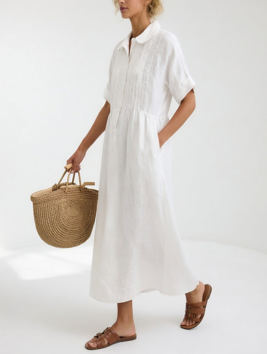 Model in the white dress seen from the side, hand in pocket, carrying a woven straw tote and wearing tan sandals. 1