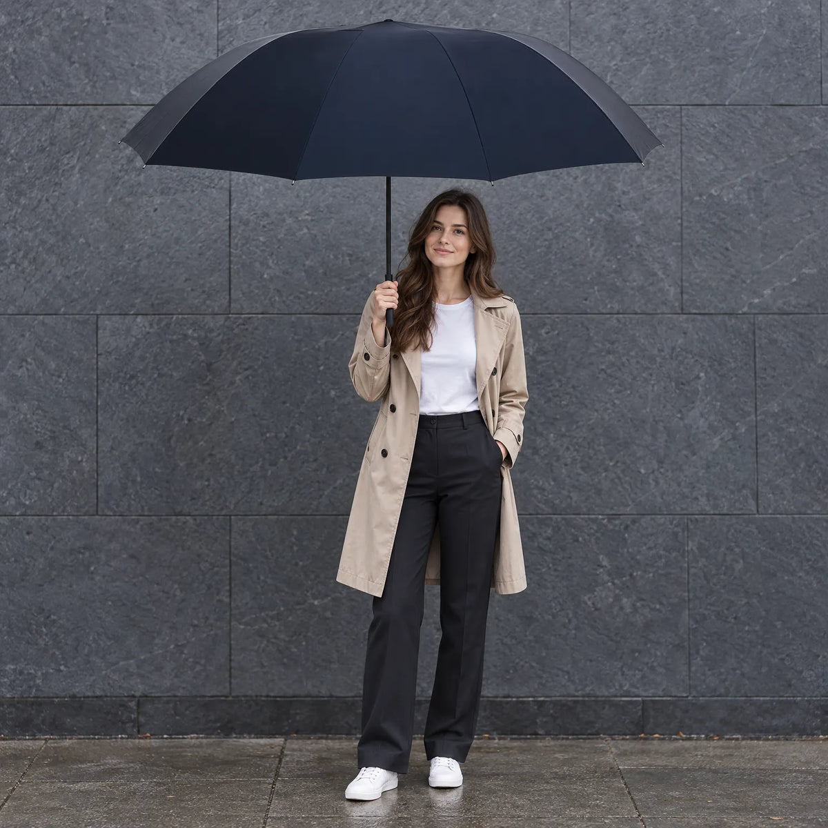 Woman in a beige trench coat and white trainers holds the large navy umbrella open against a grey stone wall. 1