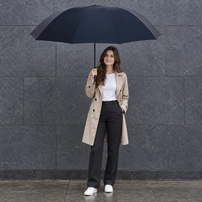 Woman in a beige trench coat and white trainers holds the large navy umbrella open against a grey stone wall. 1