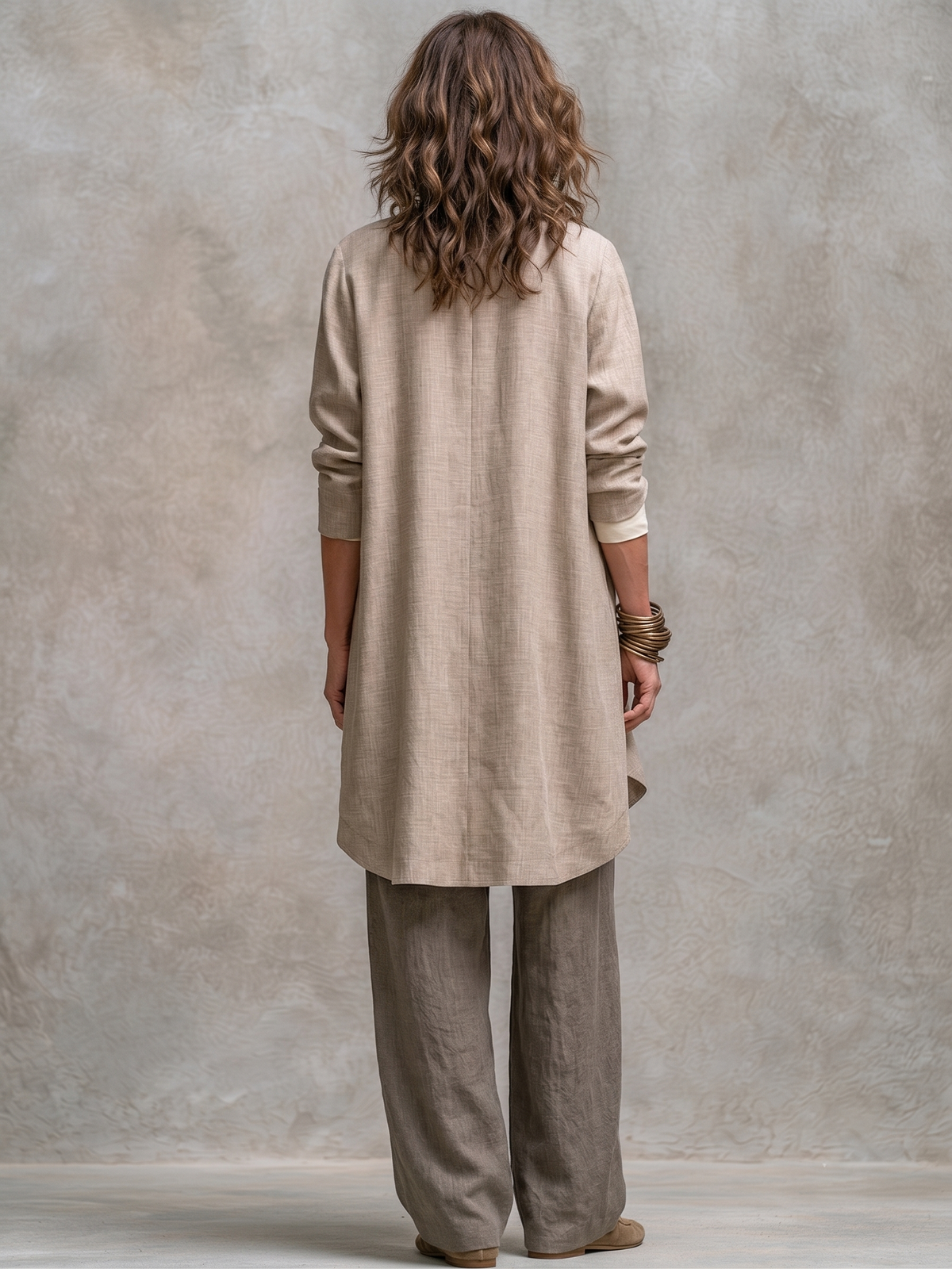 Back view of the long beige jacket with its centre seam and curved hem, worn over taupe wide-leg trousers. 6