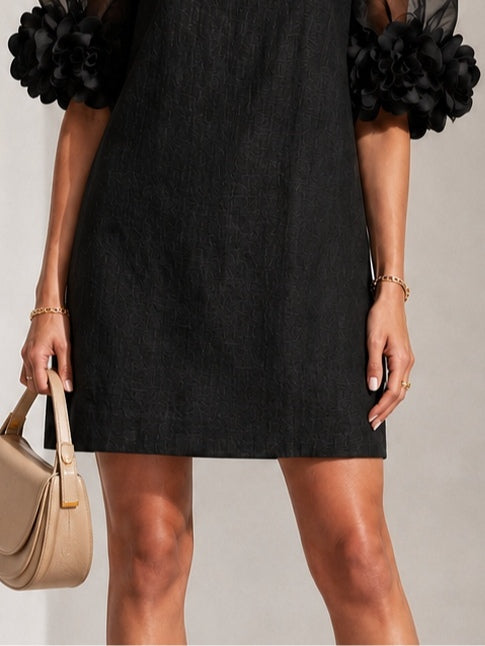 Close-up of the lower half of the black dress showing its textured fabric, straight hem and floral sleeve cuffs. 6