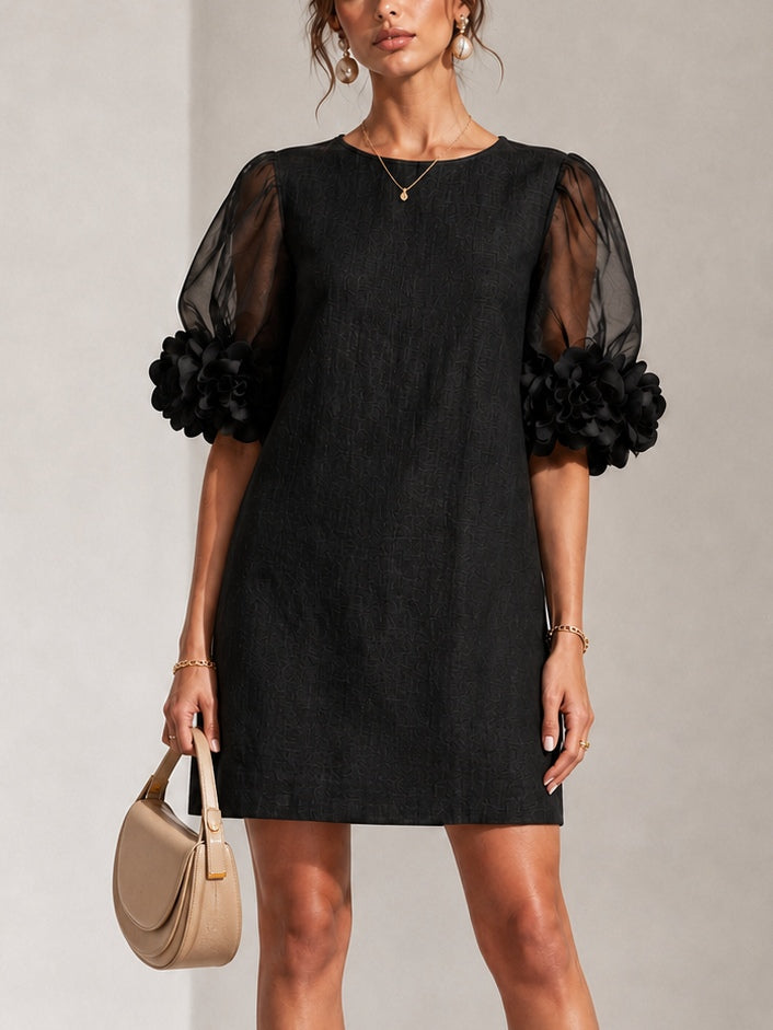 Model wearing the black textured shift dress from the front, with sheer puff sleeves, floral cuffs and a beige handbag. 1