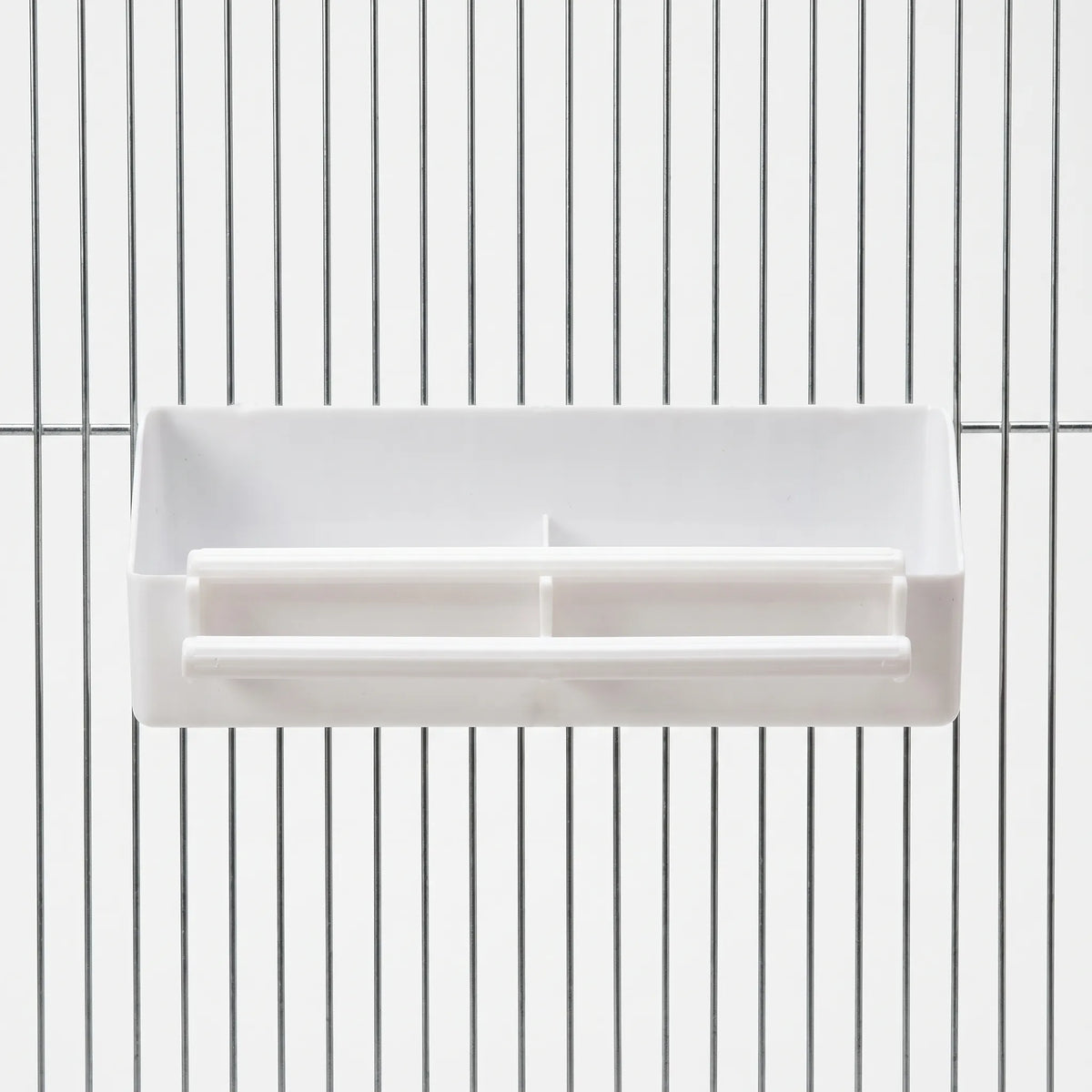 White plastic divided feeder with two perches, hooked onto the metal bars of a cage, seen straight from the front. 1