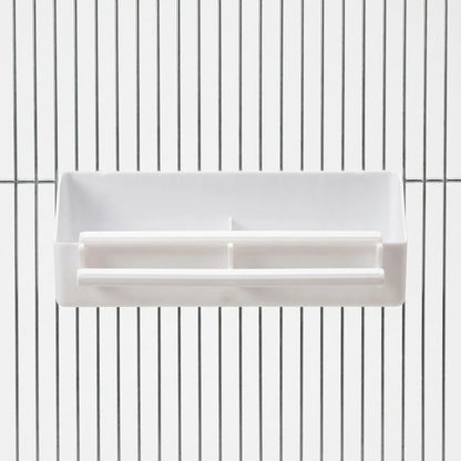 White plastic divided feeder with two perches, hooked onto the metal bars of a cage, seen straight from the front. 1