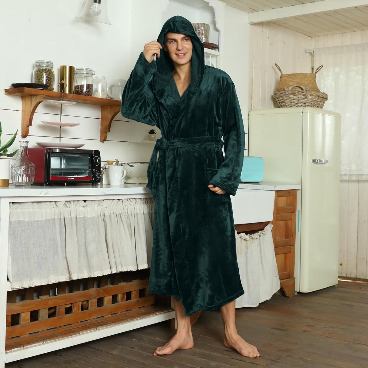 Model in the dark green hooded plush robe, hood up and belt tied, standing in a rustic kitchen 4