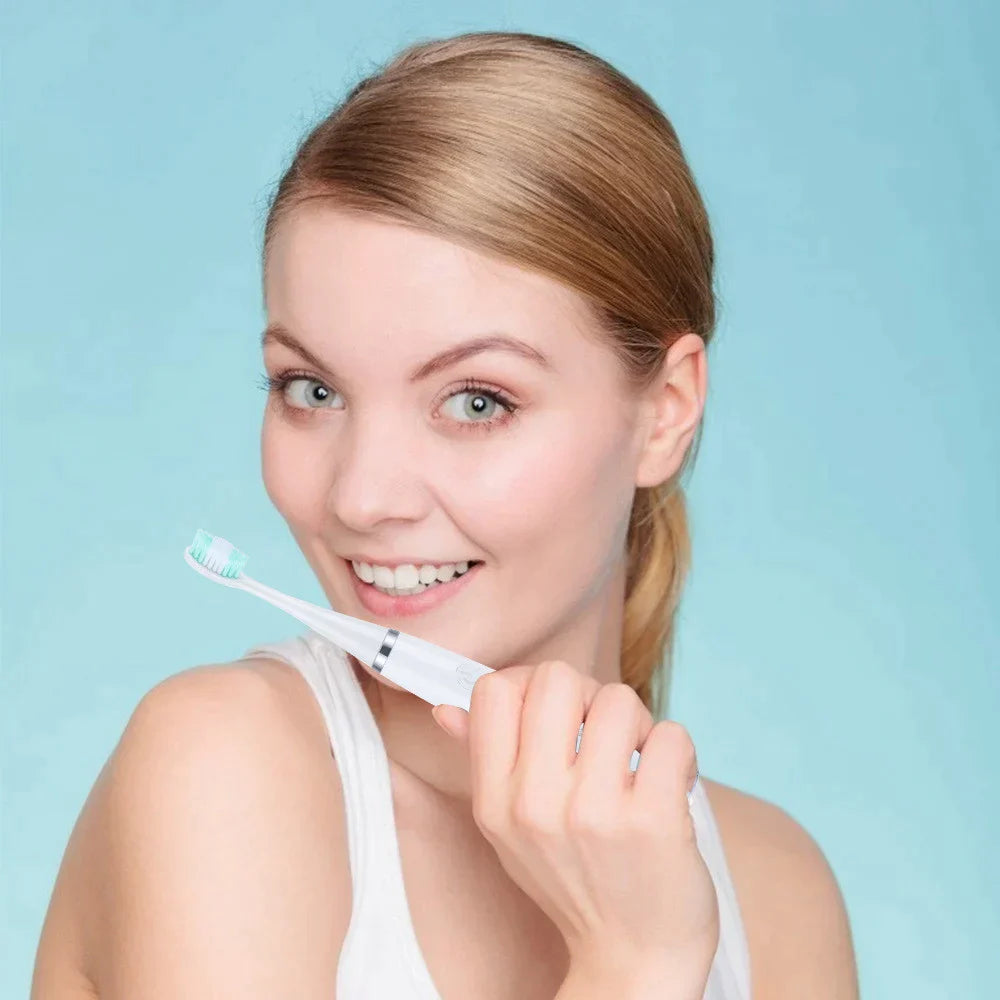 Blonde woman smiling against a pale blue background, holding the white electric toothbrush with a green-tipped brush head near her chin. 7