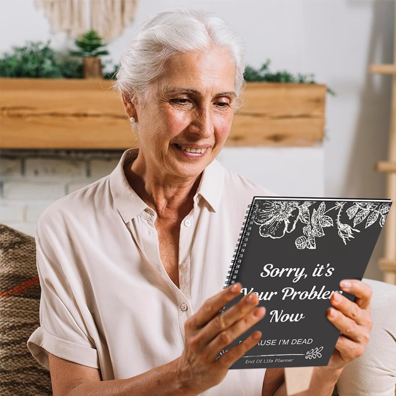 Smiling grey-haired woman in a cream blouse holds up the dark floral planner and looks at its cover. 5