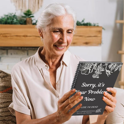 Smiling grey-haired woman in a cream blouse holds up the dark floral planner and looks at its cover. 5