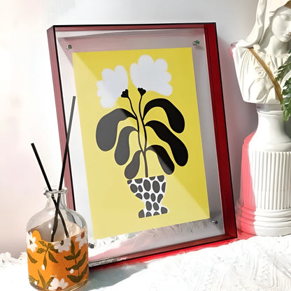 Deep red acrylic frame leaning on a white surface, holding a yellow print of a potted plant, beside a diffuser bottle and bust. 15