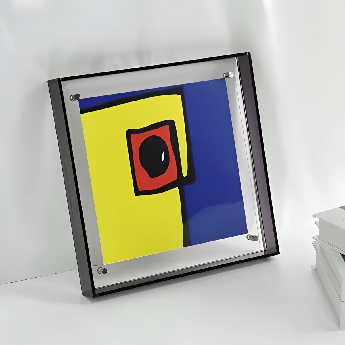 Square frame with dark edges leaning on a white surface, showing an abstract yellow, blue and red print near stacked books. 6
