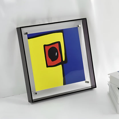 Square frame with dark edges leaning on a white surface, showing an abstract yellow, blue and red print near stacked books. 6