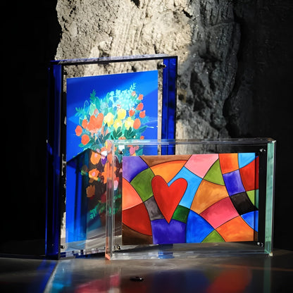Blue acrylic frame holding a flower bouquet print beside a clear landscape frame with a colourful heart pattern, against rock. 5