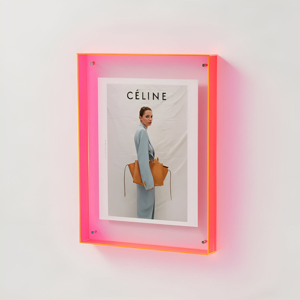 Neon pink acrylic frame mounted on a white wall, glowing softly and holding a fashion print of a woman carrying a tan bag. 18