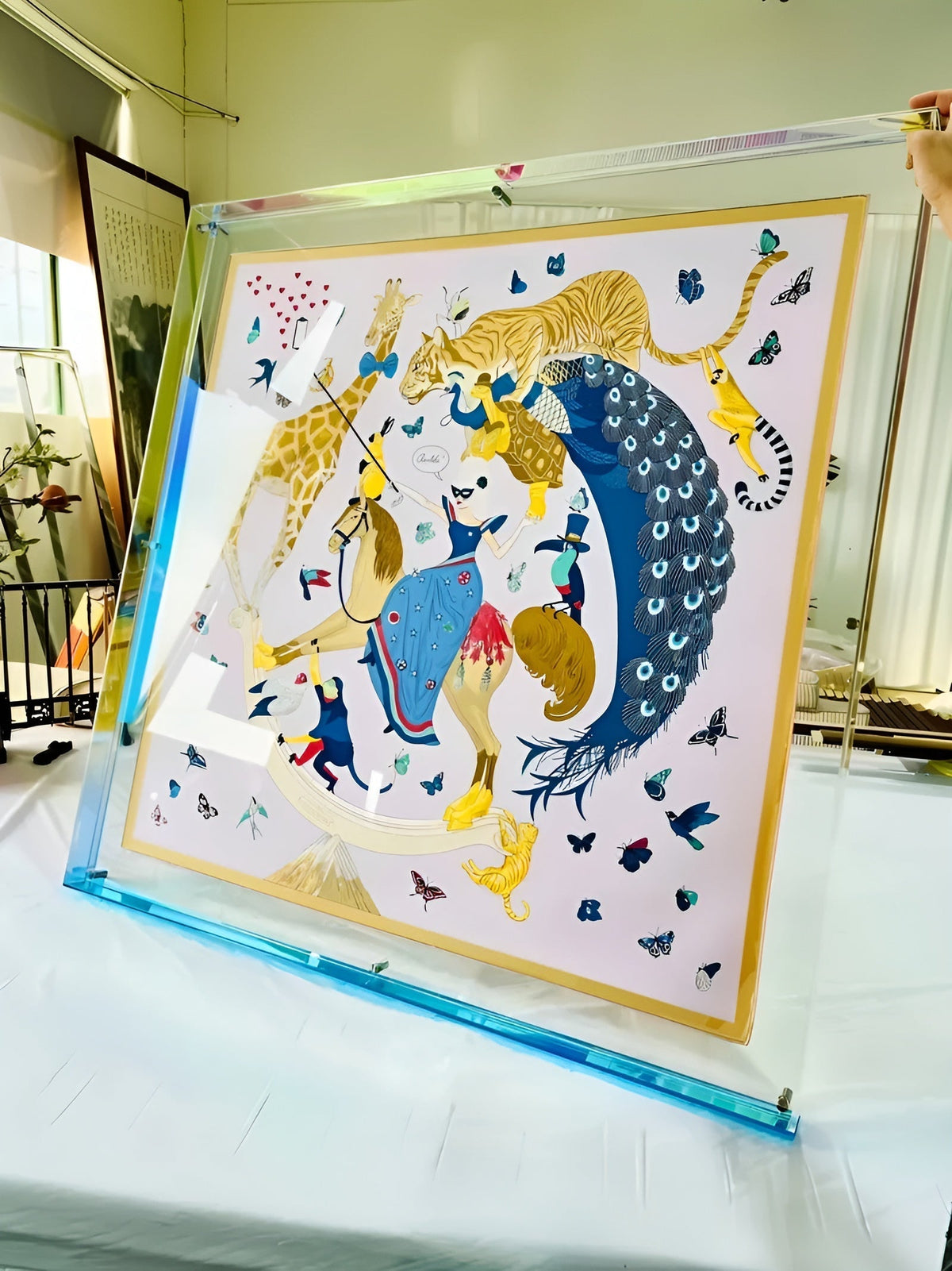 Large clear acrylic frame with a blue edge, held up by hand, displaying a silk scarf covered in animals and butterflies. 17