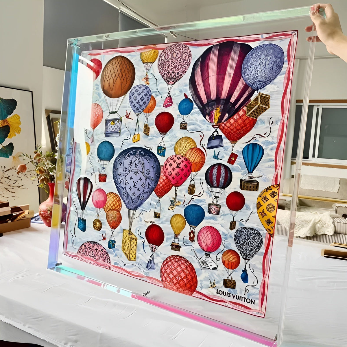Large clear acrylic frame with a rainbow-tinted edge, held by hand, displaying a silk scarf patterned with colourful hot air balloons. 11