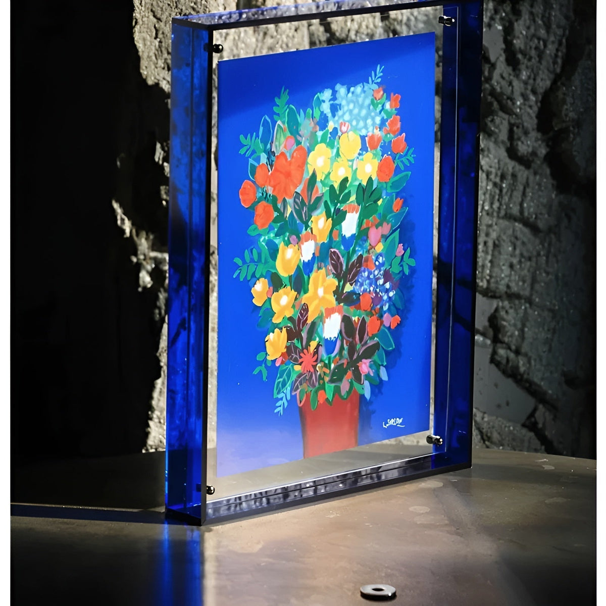 Deep blue acrylic frame standing on a metal surface against a rough stone wall, holding a bright bouquet painting. 4