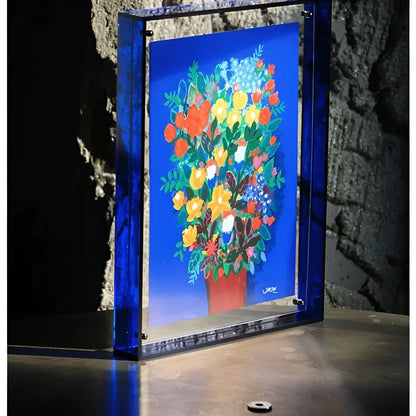 Deep blue acrylic frame standing on a metal surface against a rough stone wall, holding a bright bouquet painting. 4