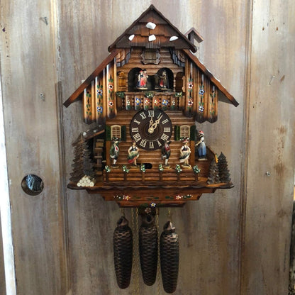 Chalet cuckoo clock hanging on a weathered wooden door, with painted flowers on the roof, dancing figures, waterwheel and three cone weights. 12