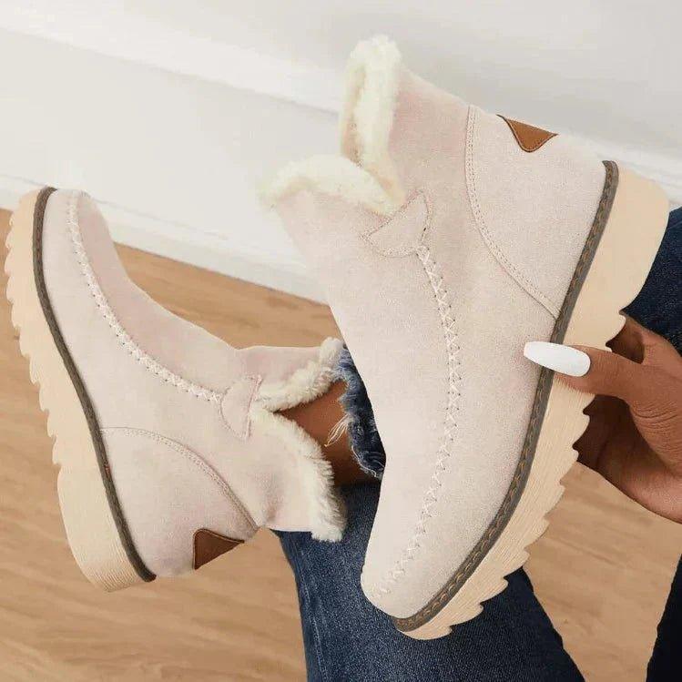Beige ankle boots held up by a model's hand, showing the stitched toe, brown heel patch and thick fleece-lined collar. 6