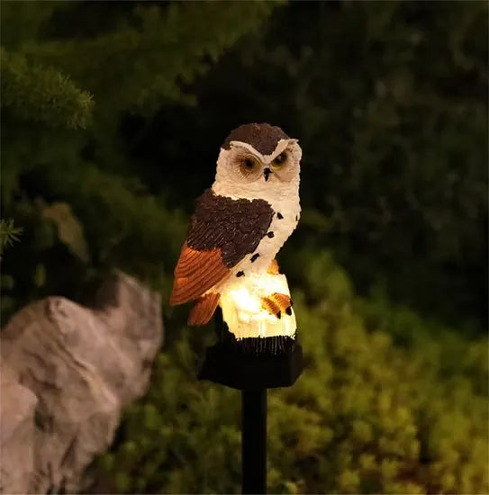 Brown-and-white owl light with russet wing tips, glowing at night on its stake beside a rock and shrubs. 7