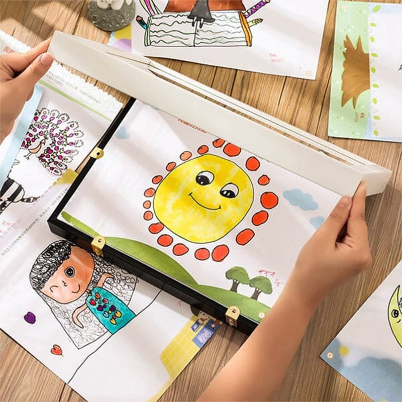 Hands lifting the cover of a black frame lying on a wooden table among a child's colourful drawings. 12