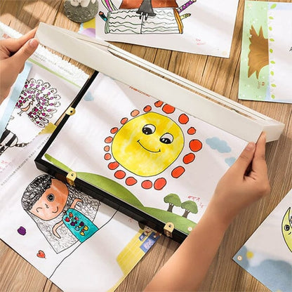 Hands lifting the cover of a black frame lying on a wooden table among a child's colourful drawings. 12