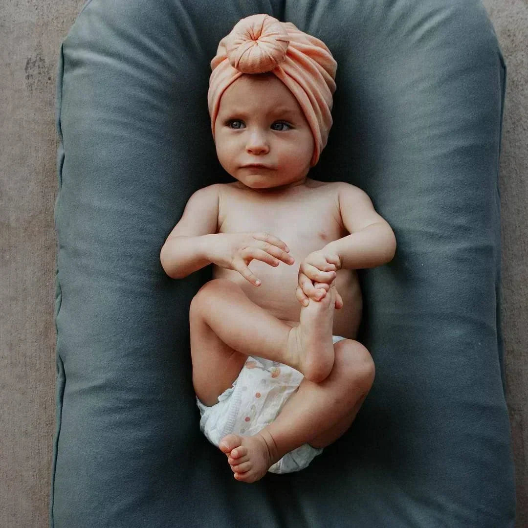 Baby in a peach knotted headwrap and nappy lying on a grey-blue lounger, viewed from above 3