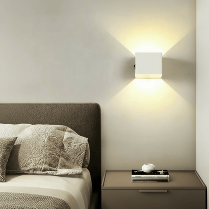 White cube wall light mounted above a bedside table beside a bed, spreading warm light up and down the wall. 3