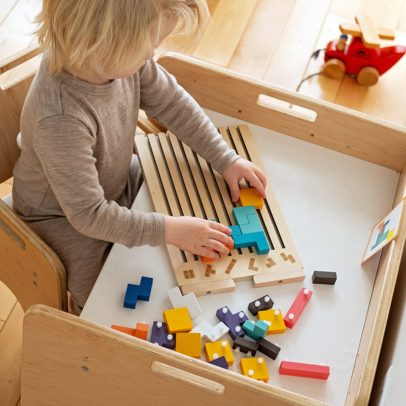 Child seated at a wooden activity table fitting teal and orange blocks into the board, with spare pegged blocks scattered nearby. 8
