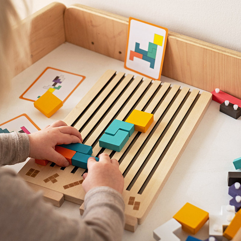 Child's hands sliding coloured blocks into the slotted board on a low table, with pattern cards propped up behind. 3