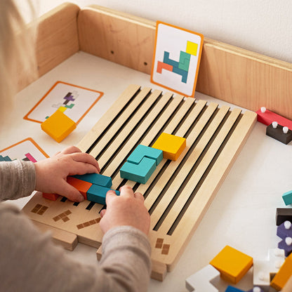 Child's hands sliding coloured blocks into the slotted board on a low table, with pattern cards propped up behind. 3