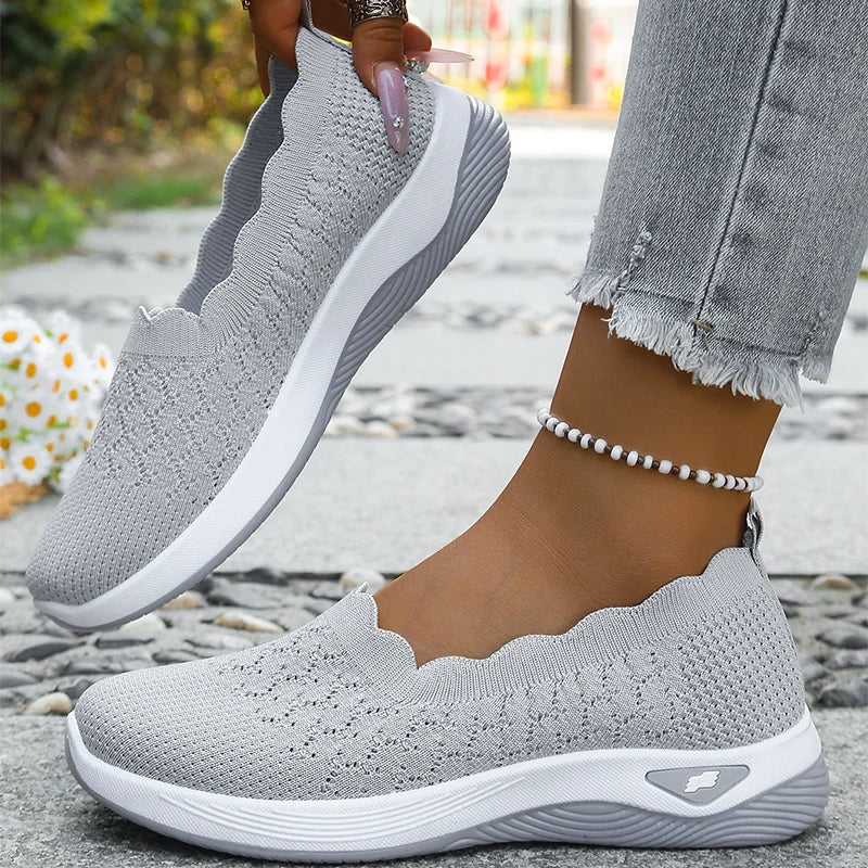 Grey knit slip-ons with white midsole, one lifted by hand and one worn, beaded anklet and daisies behind 10