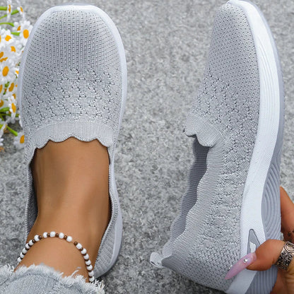 Top-down view of grey knit slip-ons showing the scalloped collar and perforated pattern, one shoe tilted by hand 11