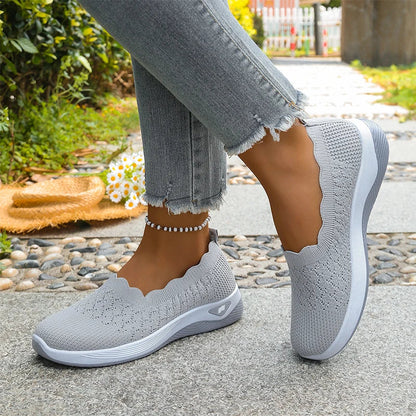 Grey knit slip-on shoes worn with frayed jeans on a paved path, daisies and a straw hat beside them 12