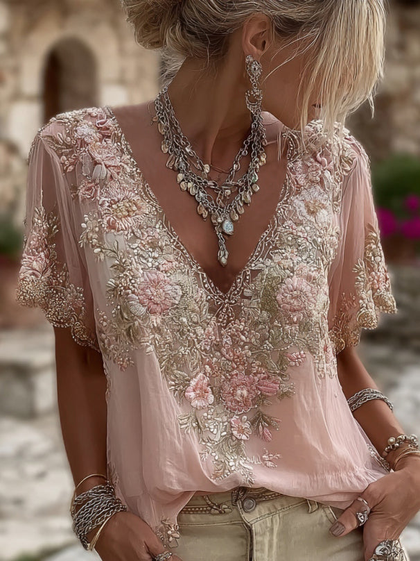 Close, portrait-style view of a pale pink short-sleeved V-neck blouse with floral embroidery, worn with layered silver necklaces and beige trousers. 1