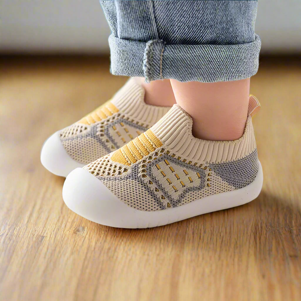 Child wearing beige and grey knit slip-on shoes with mustard stripes and white soles, standing on a wooden floor in rolled jeans. 9