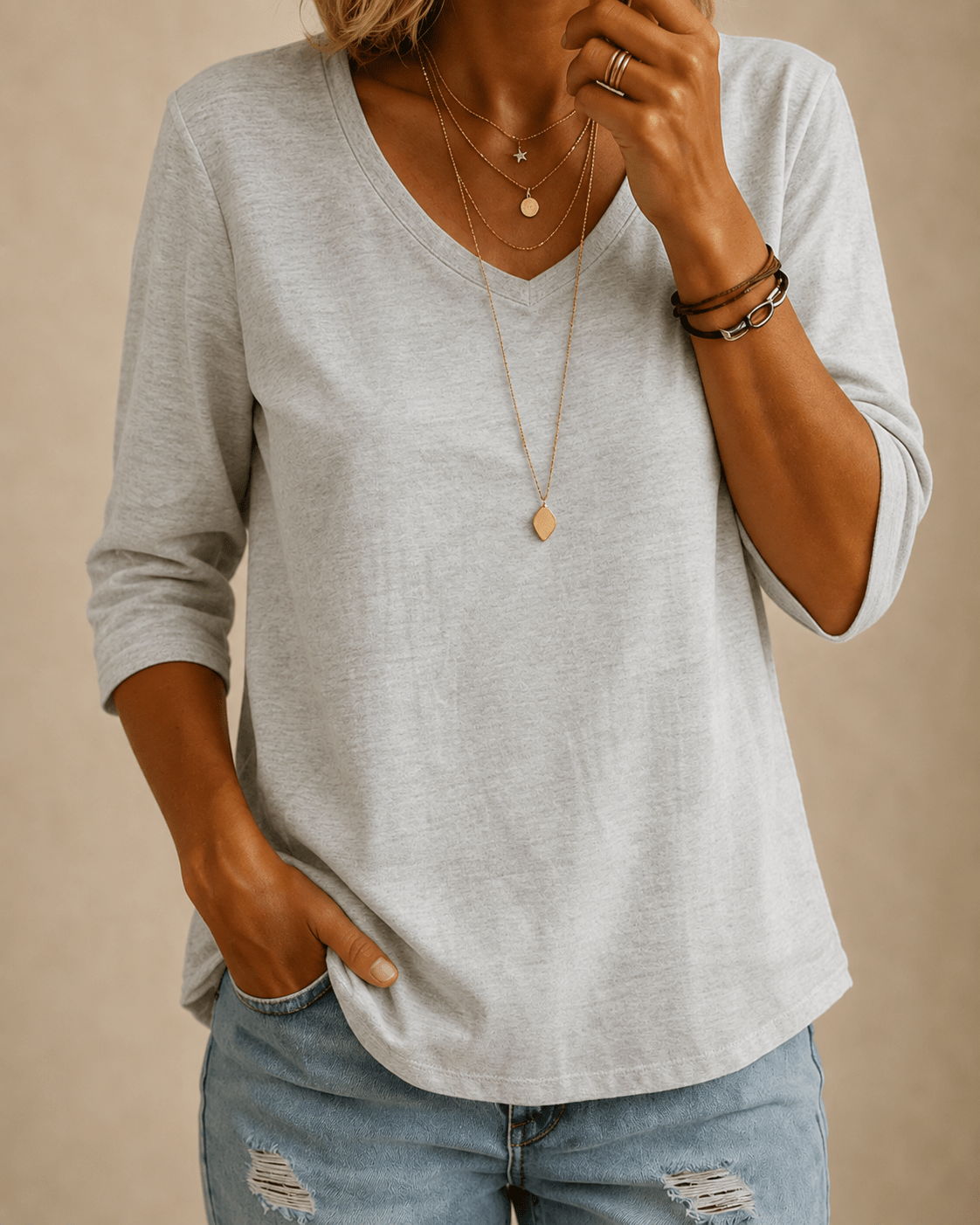 Woman wearing the light grey tee with distressed light-wash jeans, layered gold necklaces and leather bracelets, hand in 1